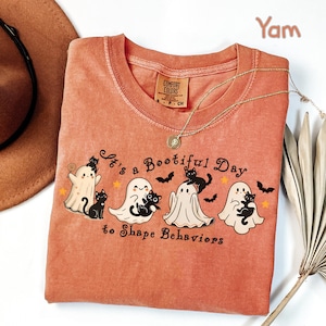 May include: A yam-colored t-shirt with a crew neck and the text "It's a Bootiful Day to Shape Behaviors." The graphic features cartoon ghosts, black cats, bats, and stars. A gold necklace is draped across the shirt.
