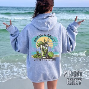 Turn up the tropical energy with our Fresh & Zesty – Minted in Miami hoodie, featuring a bold mojito graphic set against a dreamy Miami sunset beach scene. This eye-catching back print channels sunny coastal vibes, citrusy freshness, and laid-back luxury—perfect for sipping mojitos or dreaming of South Beach. A go-to for travel, lounging, or gifting anyone who loves cocktails, sunshine, and Miami heat.  Ideal gift for cocktail lovers, beachgoers, or anyone with a thing for Miami vibes.