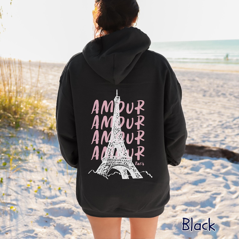 K&ouml;nnte beinhalten: Ein schwarzer Kapuzenpullover mit einer wei&szlig;en Grafik des Eiffelturms auf der R&uuml;ckseite. Das Wort AMOUR ist viermal vertikal in Hellrosa hinter dem Turm aufgedruckt, wobei das Wort Paris in kleiner wei&szlig;er Schrift nahe der Basis steht.