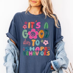May include: A navy blue t-shirt featuring the phrase "IT'S A GOOD day TO SHAPE BEHAVIORS" in bright, whimsical lettering. The design incorporates floral and heart motifs. The shirt has a relaxed, comfortable fit.