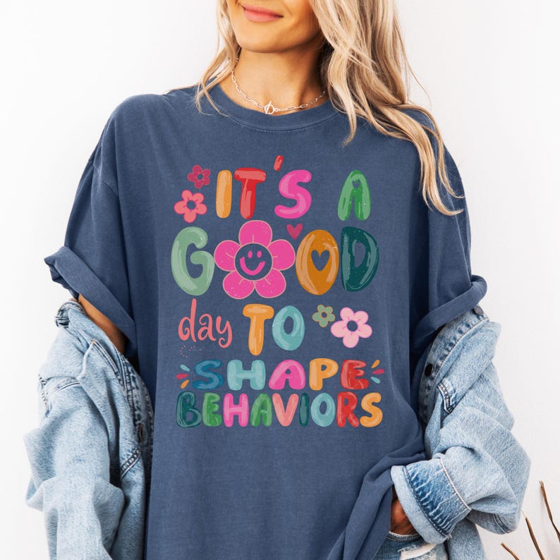 May include: A navy blue t-shirt featuring the phrase "IT'S A GOOD day TO SHAPE BEHAVIORS" in bright, whimsical lettering. The design incorporates floral and heart motifs. The shirt has a relaxed, comfortable fit.
