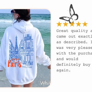 K&ouml;nnte beinhalten: Ein wei&szlig;er Kapuzenpullover mit einer blauen Strichzeichnung einer Pariser Stra&szlig;enszene inklusive Eiffelturm und dem orangefarbenen Schriftzug "see you in Paris". Die R&uuml;ckseite des Kleidungsst&uuml;cks zeigt dieses grafische Design deutlich.