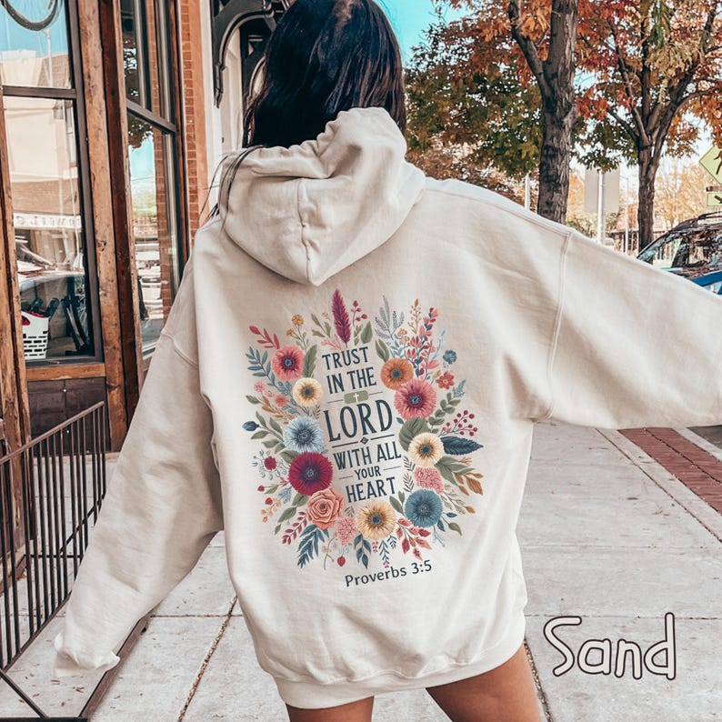 May include: Sand-colored hoodie with a floral design and the text "Trust in the Lord with all your heart" and "Proverbs 3:5". The design features colorful flowers and leaves. The hoodie has a hood and long sleeves.
