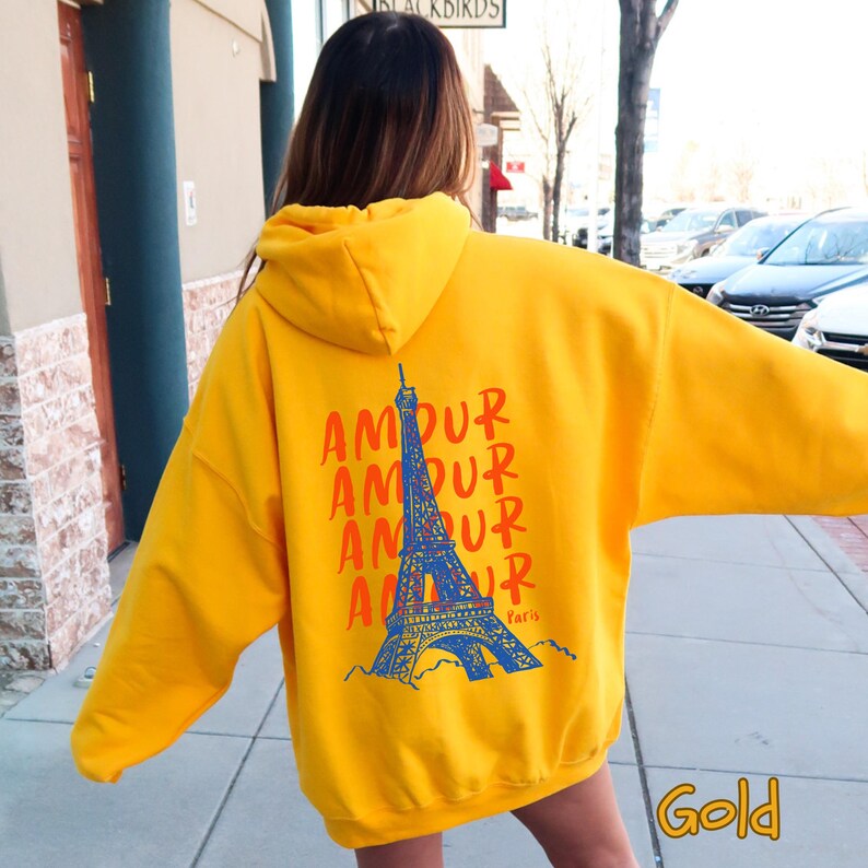K&ouml;nnte beinhalten: Ein leuchtend gelber Kapuzenpullover mit einer Grafik auf der R&uuml;ckseite. Das Design zeigt das Wort AMOUR viermal in Orange, eine blaue Illustration des Eiffelturms und das Wort Paris in kleinen blauen Buchstaben darunter gedruckt.