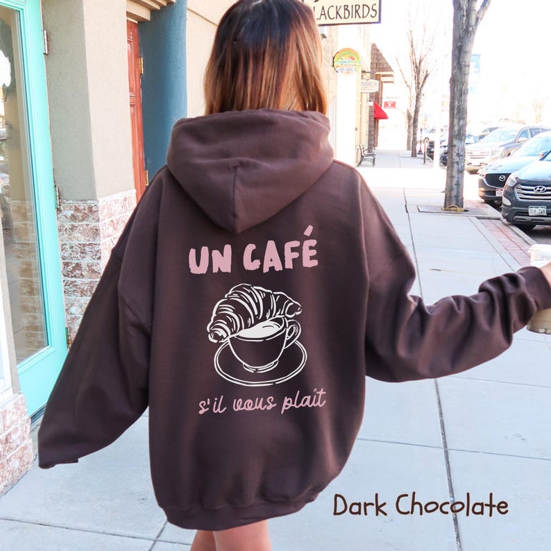 May include: A dark chocolate brown hooded sweatshirt featuring a graphic on the back. The design includes a white line drawing of a croissant resting on a coffee cup and saucer, with the text UN CAF&Eacute; and s'il vous pla&icirc;t in light pink.
