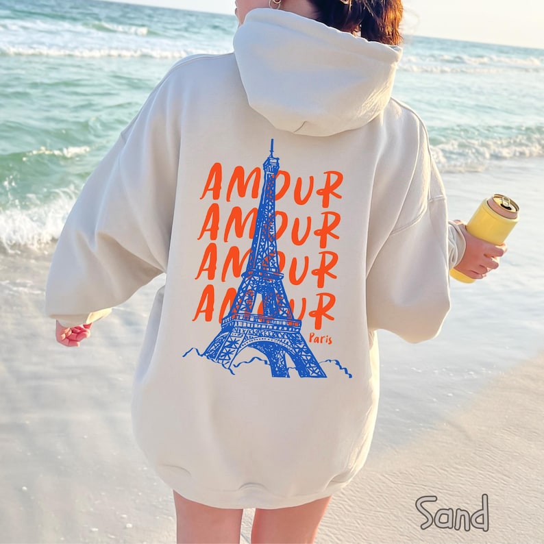 K&ouml;nnte beinhalten: Ein sandfarbener Kapuzenpullover mit einem Grafikdruck auf der R&uuml;ckseite. Das Design zeigt eine blaue Illustration des Eiffelturms, das Wort AMOUR viermal in Orange wiederholt und das Wort Paris in orangefarbener Schreibschrift.