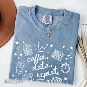 May include: Blue jean coloured t-shirt with white graphic text and illustrations. The text reads "coffee, data, repeat" with images of a coffee cup, charts, and a stopwatch. A gold necklace is draped across the shirt.