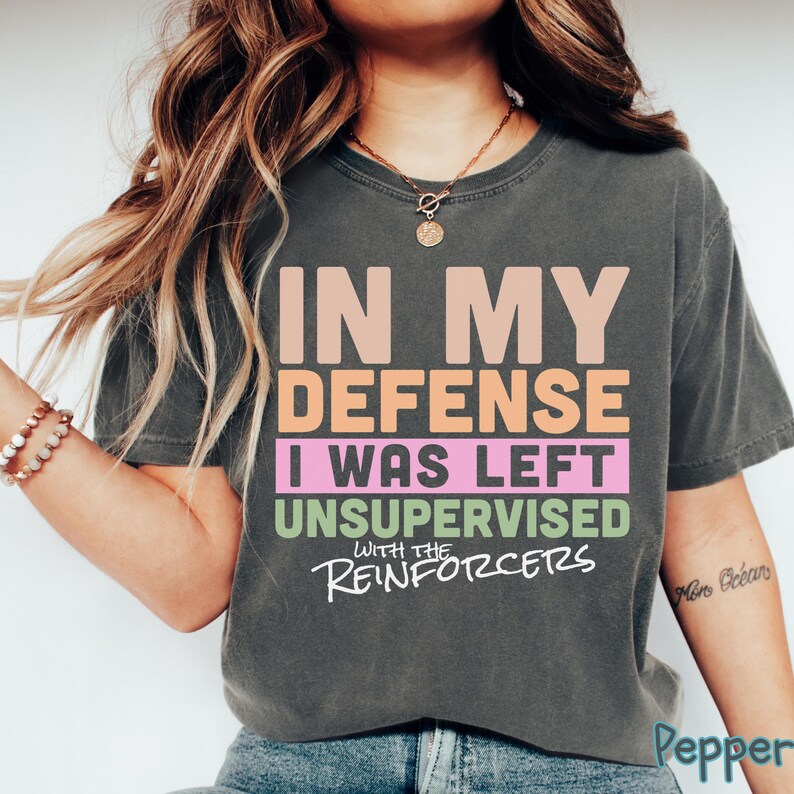 May include: Dark grey t-shirt with the slogan "IN MY DEFENSE I WAS LEFT UNSUPERVISED WITH THE REINFORCERS" in peach, pink, and green. The shirt has a crew neck and short sleeves. The text is in a bold, sans-serif font.