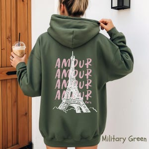 K&ouml;nnte beinhalten: Ein milit&auml;rgr&uuml;ner Kapuzenpullover mit einer wei&szlig;en Strichzeichnung des Eiffelturms auf der R&uuml;ckseite. Das Wort AMOUR ist viermal vertikal in Hellrosa neben dem Turm aufgedruckt, mit dem Wort Paris in kleiner wei&szlig;er Schrift.