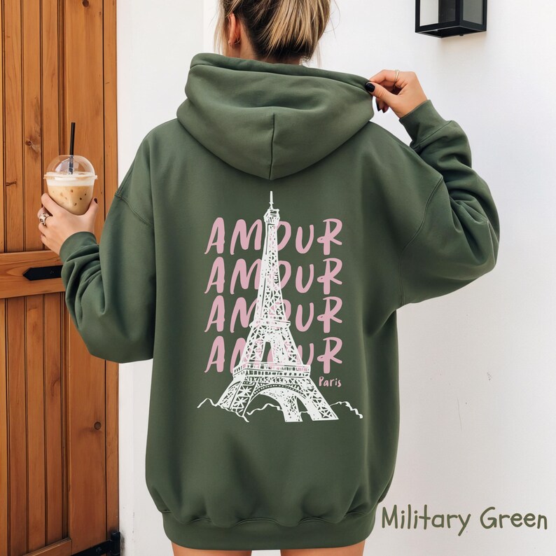 K&ouml;nnte beinhalten: Ein milit&auml;rgr&uuml;ner Kapuzenpullover mit einer wei&szlig;en Strichzeichnung des Eiffelturms auf der R&uuml;ckseite. Das Wort AMOUR ist viermal vertikal in Hellrosa neben dem Turm aufgedruckt, mit dem Wort Paris in kleiner wei&szlig;er Schrift.