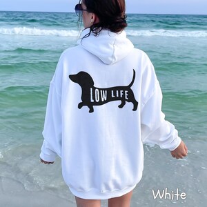 以下が含まれることがあります： 背中に黒いダックスフンドのデザインと「LOW LIFE」の文字がプリントされた白いパーカー。パーカーにはフードとゆったりとしたフィット感があります。「White」の文字が右下にプリントされています。