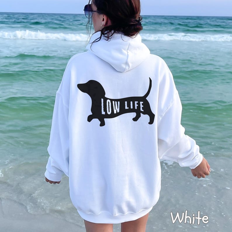 以下が含まれることがあります： 背中に黒いダックスフンドのデザインと「LOW LIFE」の文字がプリントされた白いパーカー。パーカーにはフードとゆったりとしたフィット感があります。「White」の文字が右下にプリントされています。