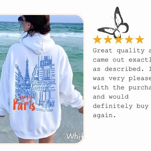 May include: A white hooded sweatshirt featuring a blue line-art illustration of a Parisian street scene with the Eiffel Tower and the text "see you in Paris" in orange. The back of the garment displays this graphic design clearly.
