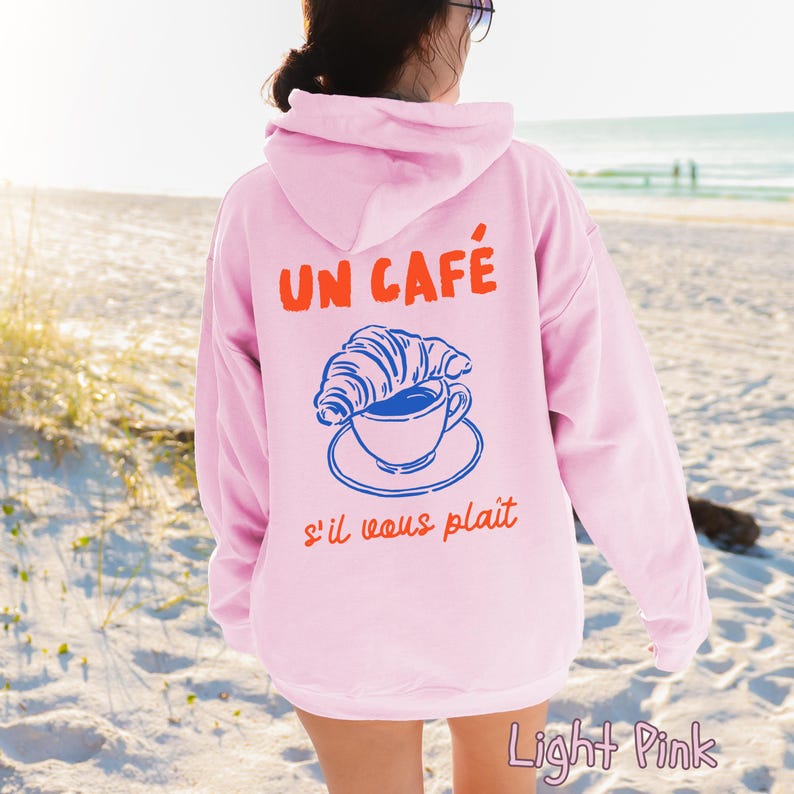 May include: A light pink hooded sweatshirt featuring a graphic on the back. The design includes the orange text UN CAF&Eacute; above a blue illustration of a croissant resting on a coffee cup and saucer, with the orange text s'il vous pla&icirc;t below.