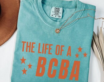 May include: A turquoise Comfort Colors T-shirt with the text "THE LIFE OF A BCBA" in orange, along with orange star graphics. A gold necklace with a pendant is draped across the shirt.