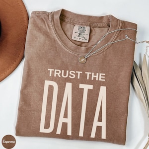 May include: Espresso-colored t-shirt with the text "TRUST THE DATA" in a bold, off-white font. The shirt has a crew neck and short sleeves. A silver necklace with a small pendant rests on the shirt. The shirt is folded on a white surface.