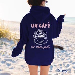 May include: A navy blue hooded sweatshirt featuring a white graphic of a croissant resting on a coffee cup and saucer. The text "UN CAF&Eacute;" appears in light pink above the graphic, and "s'il vous pla&icirc;t" is printed in light pink below the graphic.