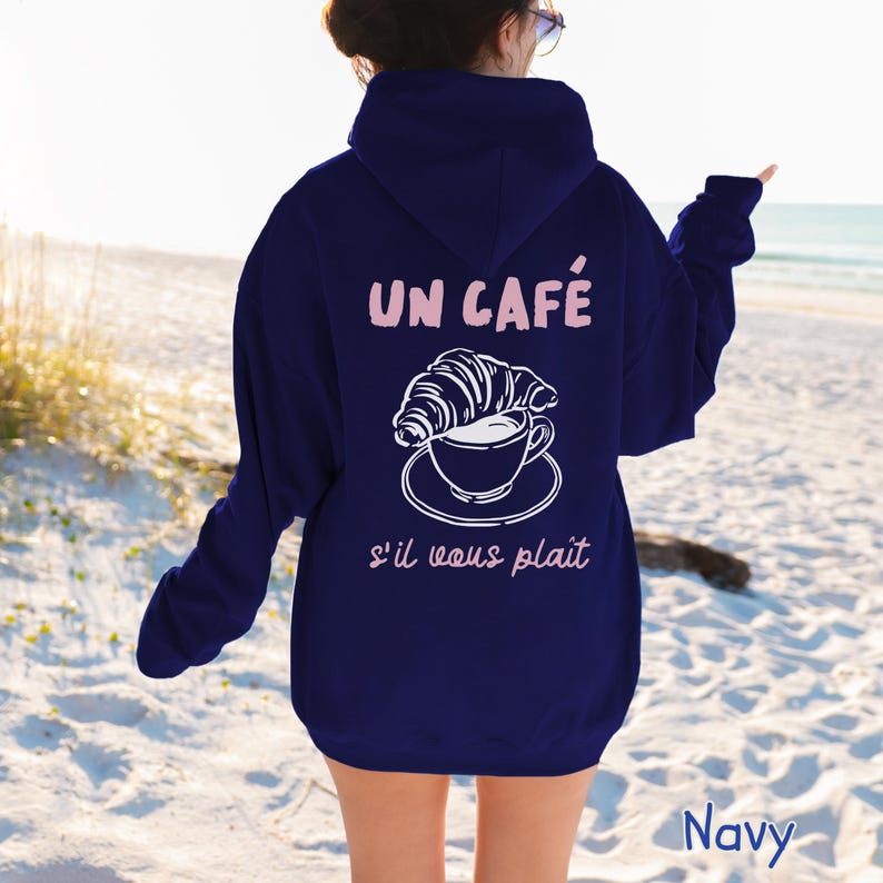 May include: A navy blue hooded sweatshirt featuring a white graphic of a croissant resting on a coffee cup and saucer. The text "UN CAF&Eacute;" appears in light pink above the graphic, and "s'il vous pla&icirc;t" is printed in light pink below the graphic.