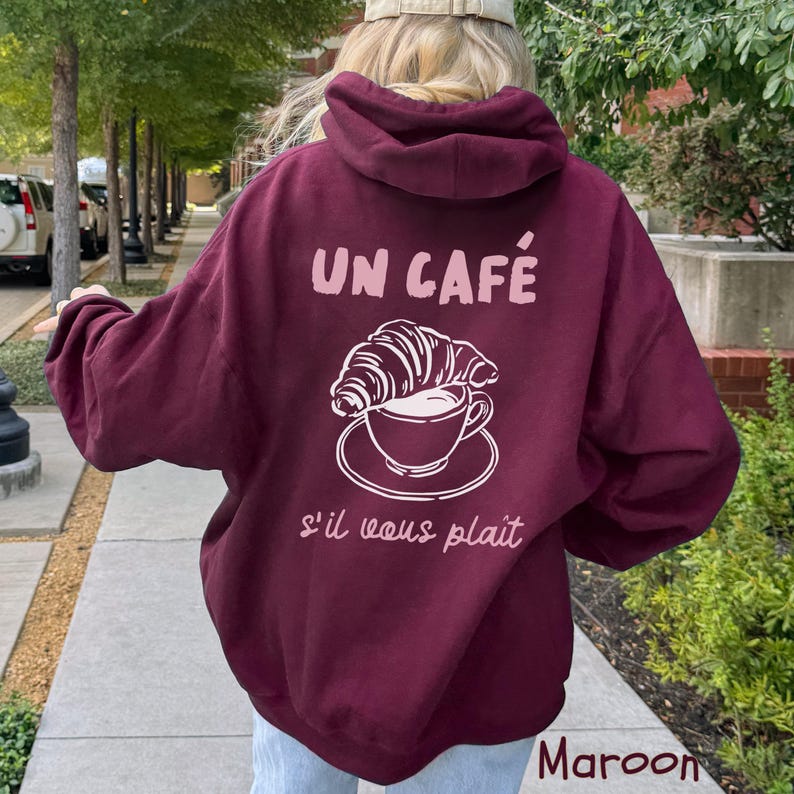 May include: A maroon hooded sweatshirt featuring a light pink graphic on the back. The graphic displays a coffee cup with a croissant resting on top, accompanied by the text "UN CAF&Eacute;" above and "s'il vous pla&icirc;t" below in a script font.