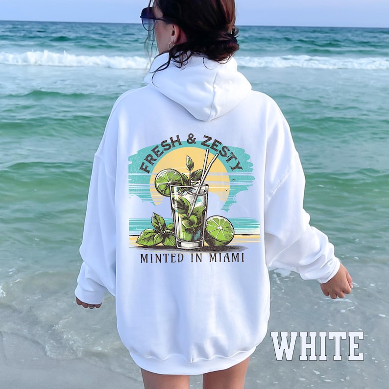 Turn up the tropical energy with our Fresh & Zesty – Minted in Miami hoodie, featuring a bold mojito graphic set against a dreamy Miami sunset beach scene. This eye-catching back print channels sunny coastal vibes, citrusy freshness, and laid-back luxury—perfect for sipping mojitos or dreaming of South Beach. A go-to for travel, lounging, or gifting anyone who loves cocktails, sunshine, and Miami heat.  Ideal gift for cocktail lovers, beachgoers, or anyone with a thing for Miami vibes.