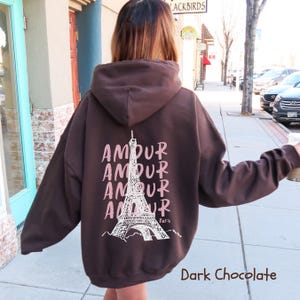 K&ouml;nnte beinhalten: Ein dunkel schokoladenbrauner Kapuzenpullover mit einem wei&szlig;en Grafikdruck auf der R&uuml;ckseite. Das Design zeigt den Eiffelturm, zentriert zwischen vier vertikal &uuml;bereinander angeordneten Zeilen des Wortes AMOUR, mit dem Wort Paris darunter.