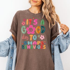 May include: A brown t-shirt featuring the phrase "IT'S A GOOD day TO SHAPE BEHAVIORS" in bright, whimsical lettering. The design incorporates floral and star motifs. The shirt has a relaxed, comfortable fit.