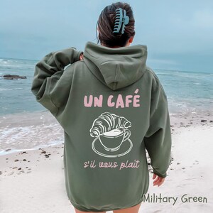 May include: An olive green hooded sweatshirt featuring a graphic on the back. The design includes a white line drawing of a croissant resting on a coffee cup and saucer, with the pink text "UN CAF&Eacute;" above and "s'il vous pla&icirc;t" below.