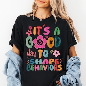 May include: Black t-shirt featuring the phrase "IT'S A GOOD day TO SHAPE BEHAVIORS" in bright, cheerful colours. The design incorporates flowers, hearts, and a smiling flower. The word "black" is printed at the bottom right.