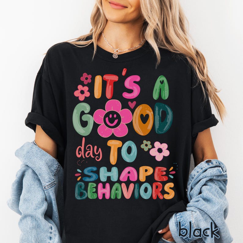 May include: Black t-shirt featuring the phrase "IT'S A GOOD day TO SHAPE BEHAVIORS" in bright, cheerful colours. The design incorporates flowers, hearts, and a smiling flower. The word "black" is printed at the bottom right.