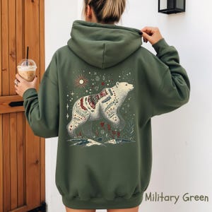 May include: Military green hoodie with a white bear illustration on the back, adorned with detailed patterns and floral elements. The design includes a sun and stars. The hoodie has a drawstring hood and long sleeves.