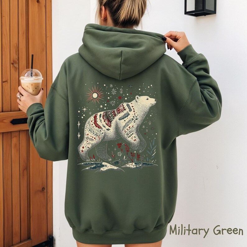 May include: Military green hoodie with a white bear illustration on the back, adorned with detailed patterns and floral elements. The design includes a sun and stars. The hoodie has a drawstring hood and long sleeves.