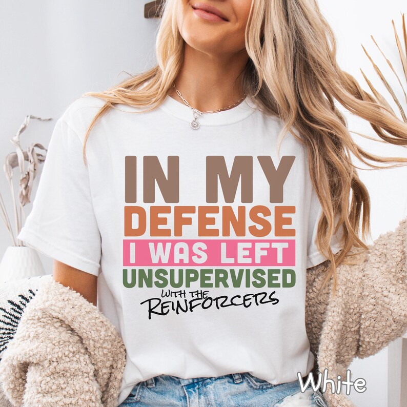 May include: White t-shirt featuring the text "IN MY DEFENSE I WAS LEFT UNSUPERVISED WITH THE REINFORCERS" in brown, orange, pink, green, and black. A casual style t-shirt.