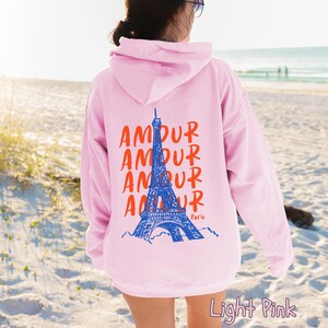 K&ouml;nnte beinhalten: Ein hellrosa Kapuzenpullover mit einer Grafik auf der R&uuml;ckseite. Das Design zeigt eine blaue Illustration des Eiffelturms, das Wort AMOUR viermal in Orange wiederholt und das Wort Paris in kleiner oranger Schrift unter dem Turm.
