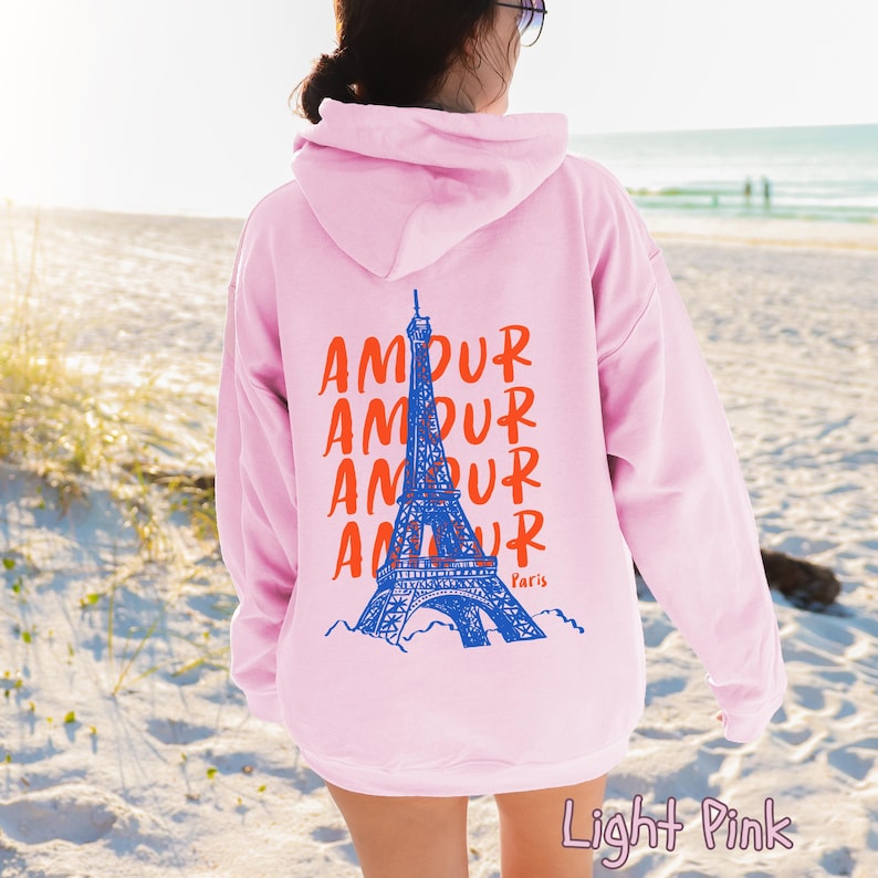 K&ouml;nnte beinhalten: Ein hellrosa Kapuzenpullover mit einer Grafik auf der R&uuml;ckseite. Das Design zeigt eine blaue Illustration des Eiffelturms, das Wort AMOUR viermal in Orange wiederholt und das Wort Paris in kleiner oranger Schrift unter dem Turm.