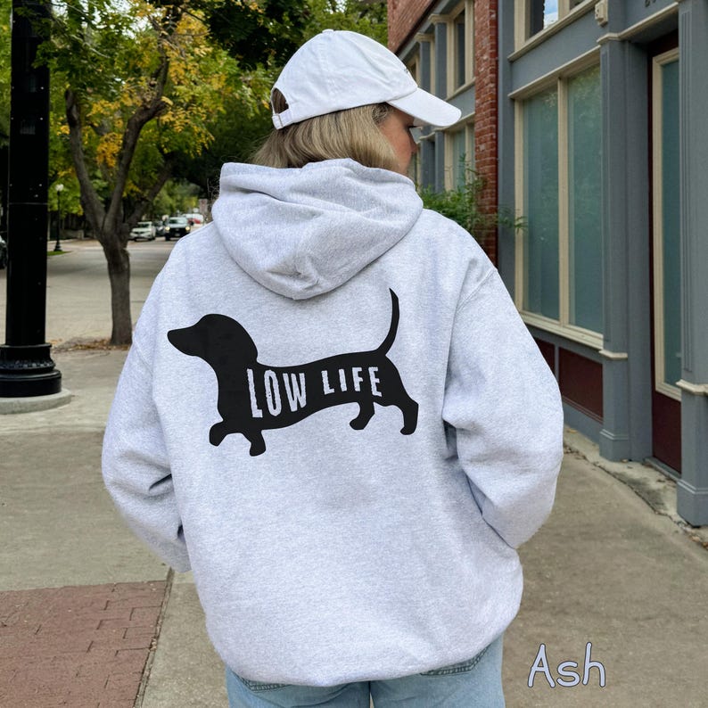 以下が含まれることがあります： 黒いダックスフントのグラフィックと犬の体に「LOW LIFE」の文字がプリントされた、明るいグレーのパーカー。フード付きで、柔らかい素材で作られています。モデルは白い野球帽をかぶっています。