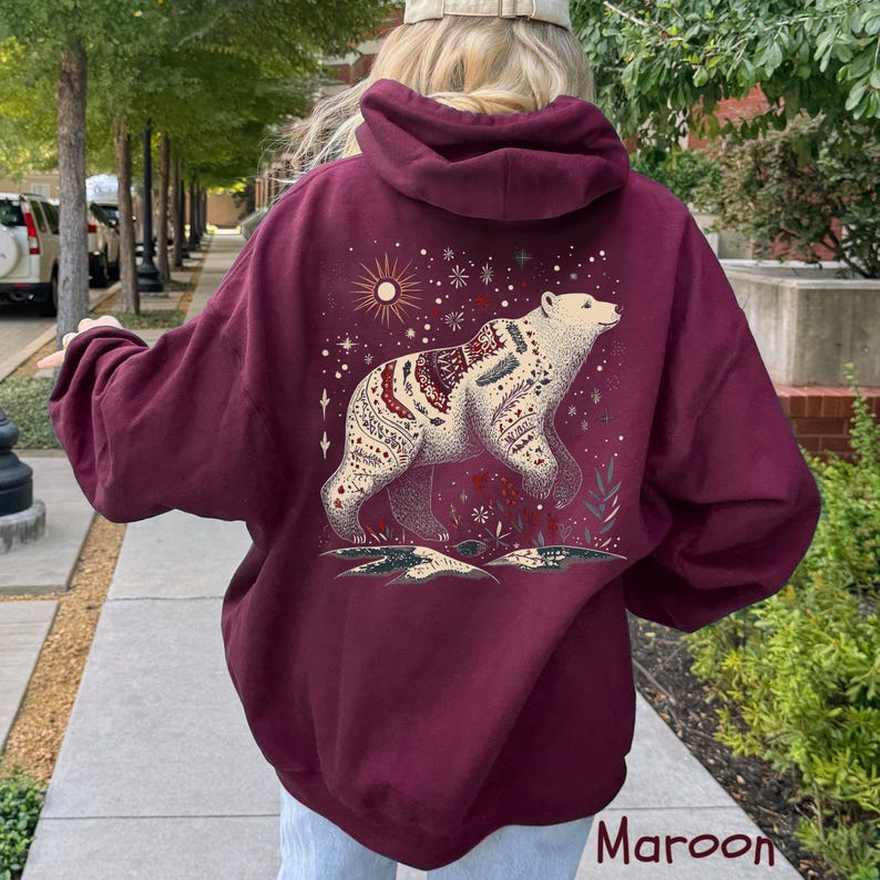 May include: Maroon coloured hoodie with a detailed illustration of a bear. The bear is white with red and cream patterns, set against a backdrop of stars and a sun. The word "Maroon" is printed at the bottom of the image.