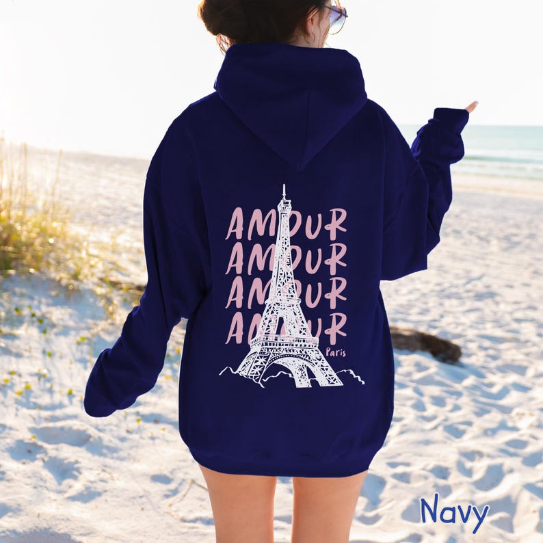 K&ouml;nnte beinhalten: Ein marineblauer Kapuzenpullover mit einer wei&szlig;en Grafik des Eiffelturms auf der R&uuml;ckseite. Das Wort AMOUR ist viermal vertikal in Hellrosa hinter dem Turm aufgedruckt, wobei das Wort Paris in kleinen wei&szlig;en Buchstaben darunter steht.