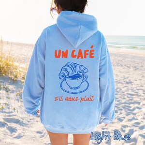 May include: A light blue hooded sweatshirt featuring a graphic on the back. The design includes the orange text UN CAF&Eacute; and s'il vous pla&icirc;t, with a blue line drawing of a croissant resting on a coffee cup and saucer in the center of the back.