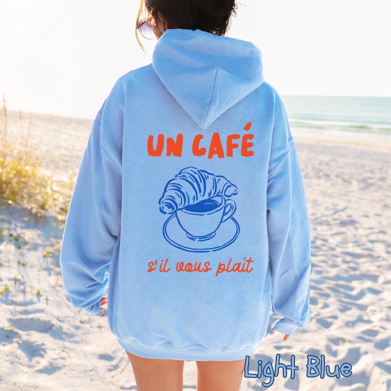 May include: A light blue hooded sweatshirt featuring a graphic on the back. The design includes the orange text UN CAF&Eacute; and s'il vous pla&icirc;t, with a blue line drawing of a croissant resting on a coffee cup and saucer in the center of the back.