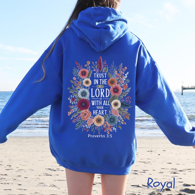 Puede incluir: Sudadera con capucha azul real con un dise&ntilde;o floral y el texto "Trust in the Lord with all your heart" y "Proverbios 3:5". El dise&ntilde;o presenta flores y follaje coloridos. La sudadera con capucha se muestra en una persona en la playa.