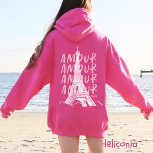 K&ouml;nnte beinhalten: Ein leuchtend pinkfarbener Kapuzenpullover mit einer wei&szlig;en Grafik auf der R&uuml;ckseite. Das Design zeigt den Eiffelturm, hinter dem das Wort AMOUR viermal untereinander steht, sowie das Wort Paris in kleiner Schreibschrift darunter.