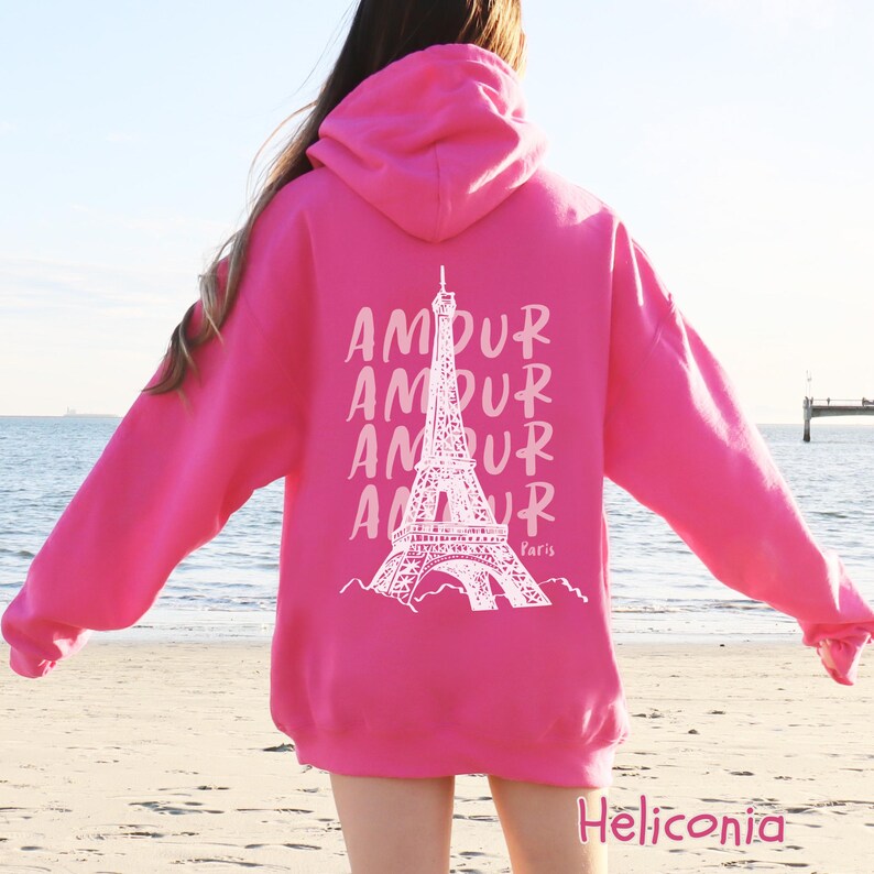 K&ouml;nnte beinhalten: Ein leuchtend pinkfarbener Kapuzenpullover mit einer wei&szlig;en Grafik auf der R&uuml;ckseite. Das Design zeigt den Eiffelturm, hinter dem das Wort AMOUR viermal untereinander steht, sowie das Wort Paris in kleiner Schreibschrift darunter.