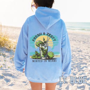 Turn up the tropical energy with our Fresh & Zesty – Minted in Miami hoodie, featuring a bold mojito graphic set against a dreamy Miami sunset beach scene. This eye-catching back print channels sunny coastal vibes, citrusy freshness, and laid-back luxury—perfect for sipping mojitos or dreaming of South Beach. A go-to for travel, lounging, or gifting anyone who loves cocktails, sunshine, and Miami heat.  Ideal gift for cocktail lovers, beachgoers, or anyone with a thing for Miami vibes.