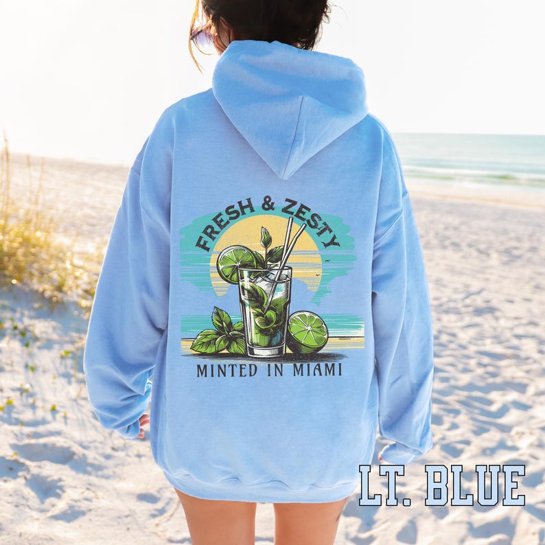 Turn up the tropical energy with our Fresh & Zesty – Minted in Miami hoodie, featuring a bold mojito graphic set against a dreamy Miami sunset beach scene. This eye-catching back print channels sunny coastal vibes, citrusy freshness, and laid-back luxury—perfect for sipping mojitos or dreaming of South Beach. A go-to for travel, lounging, or gifting anyone who loves cocktails, sunshine, and Miami heat.  Ideal gift for cocktail lovers, beachgoers, or anyone with a thing for Miami vibes.