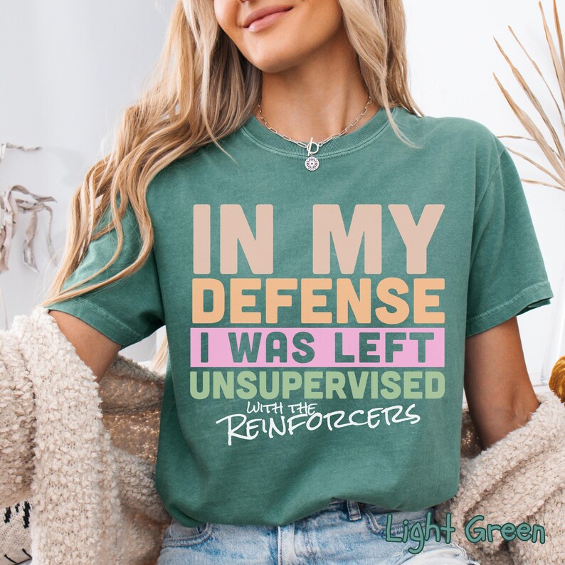 May include: Light green t-shirt featuring the text "IN MY DEFENSE I WAS LEFT UNSUPERVISED WITH THE REINFORCERS" in multiple colours. The shirt has a crew neck and short sleeves.