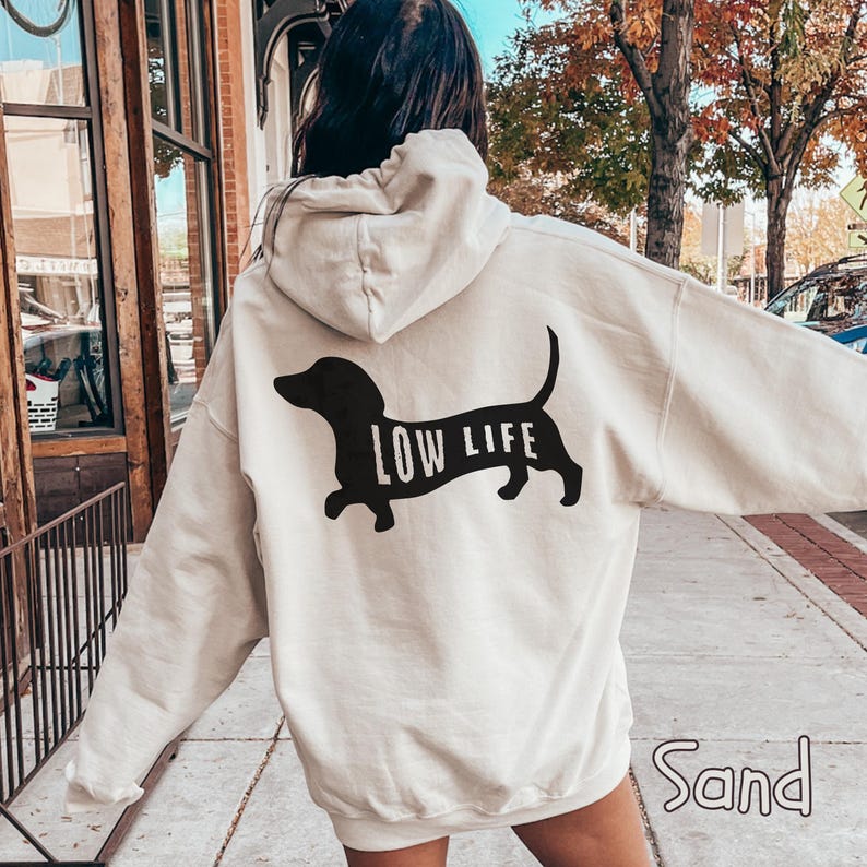 May include: Sand-colored hoodie with a black dachshund dog graphic and the words "LOW LIFE". The hoodie has a drawstring hood and long sleeves. The word "Sand" is printed at the bottom of the image.