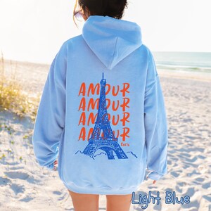 K&ouml;nnte beinhalten: Ein hellblauer Kapuzenpullover mit einer dunkelblauen Grafik des Eiffelturms auf der R&uuml;ckseite. Das Wort AMOUR ist viermal vertikal in Orange &uuml;ber den Turm gedruckt, wobei das Wort Paris in kleiner oranger Schreibschrift darunter steht.