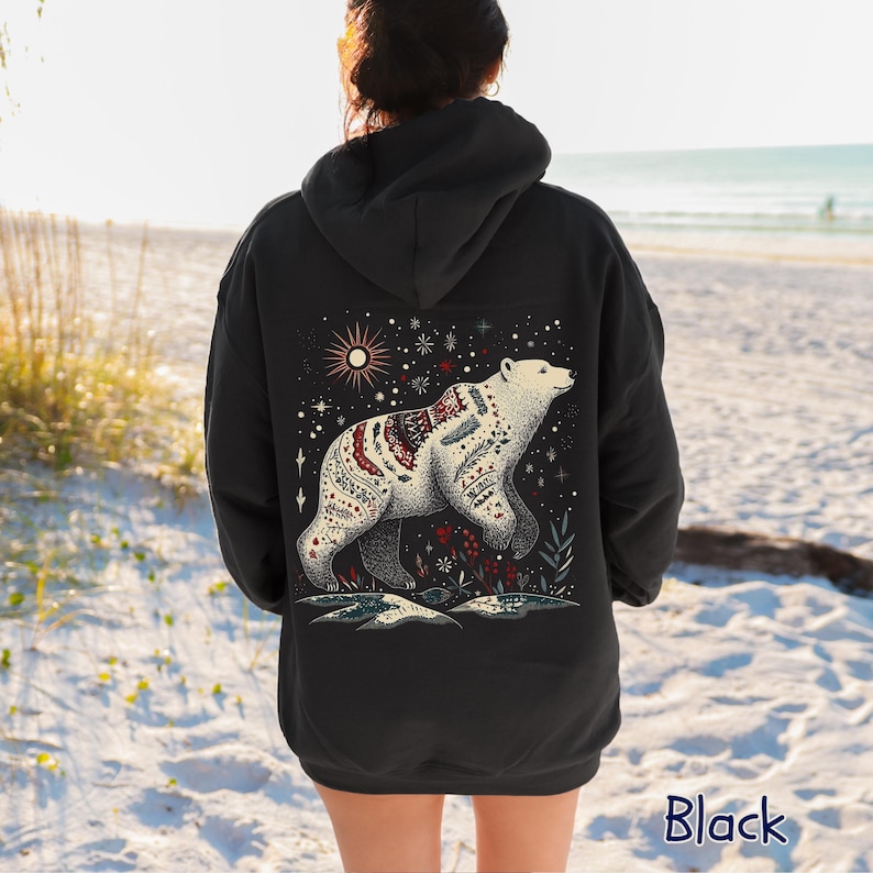 May include: Black hoodie with a detailed illustration of a polar bear adorned with intricate patterns. The design includes a starry sky, sun, and floral elements. The word "Black" is printed at the bottom of the hoodie.