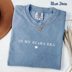 May include: A blue jean colored t-shirt with the text "IN MY BCABA ERA" in white, along with a small white heart. A gold necklace with a pendant is draped across the shirt. The shirt is a Comfort Colors brand.