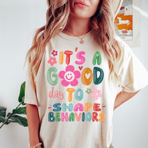 May include: A cream-coloured t-shirt featuring the phrase "It's a Good Day to Shape Behaviors" in bright, playful lettering. The design incorporates flower and heart motifs, giving a cheerful, educational feel. The t-shirt is a relaxed, comfortable style.